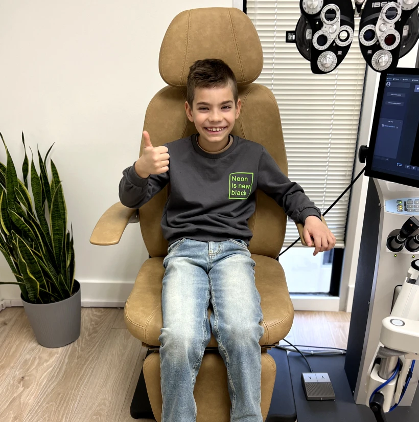 Young pediatric patient seated in exam chair smiling with thumbs up after orthokeratology fitting